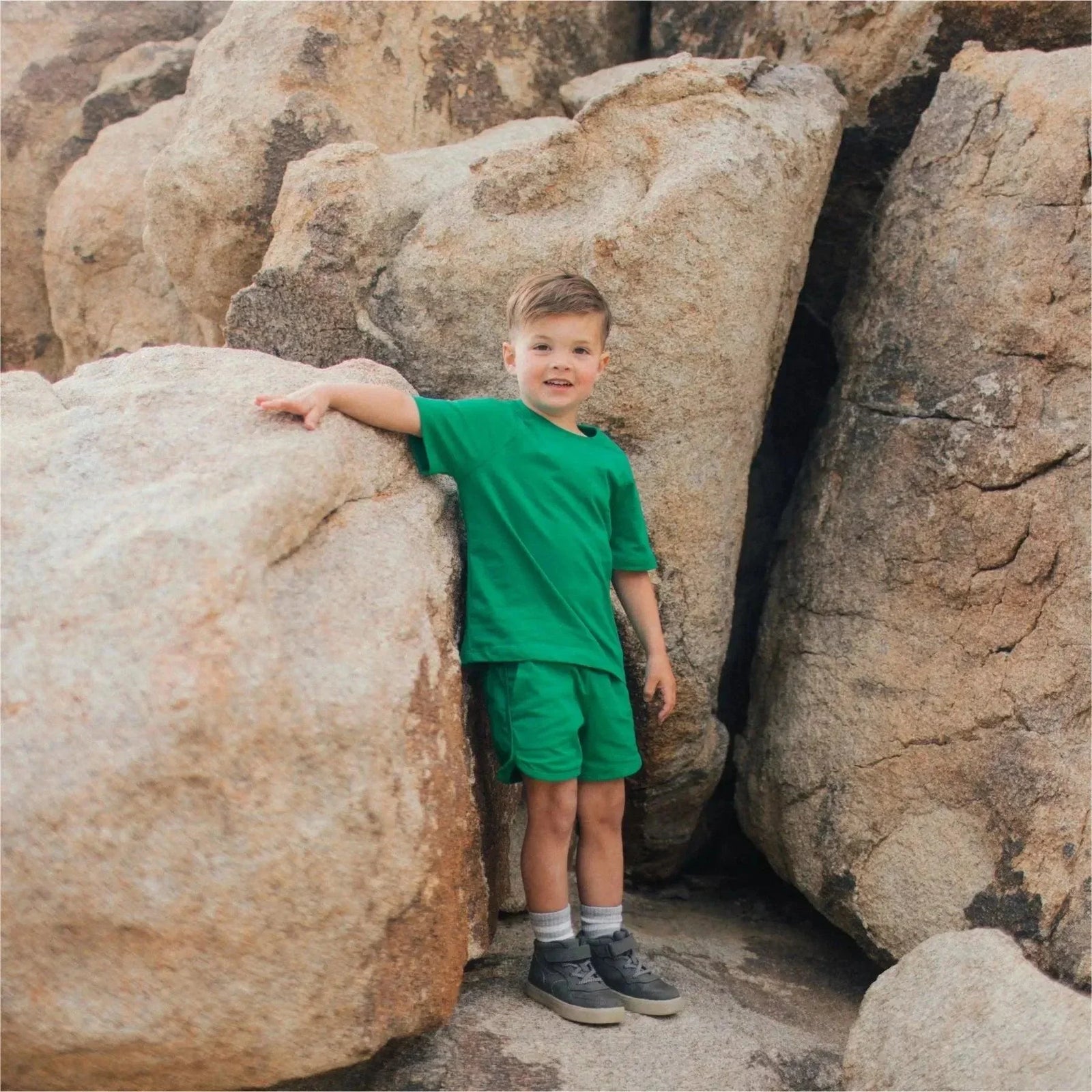 Young boy in green Jackalo activewear stands among large rocks outdoors.