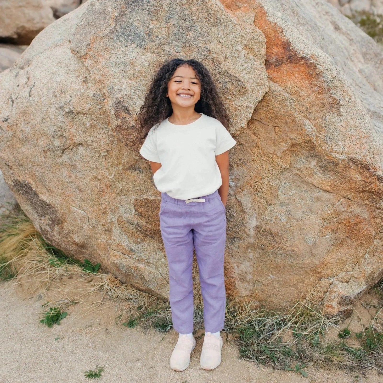 Smiling child wearing Jackalo sustainable kids clothes, white t-shirt, purple pants, outdoors by boulders