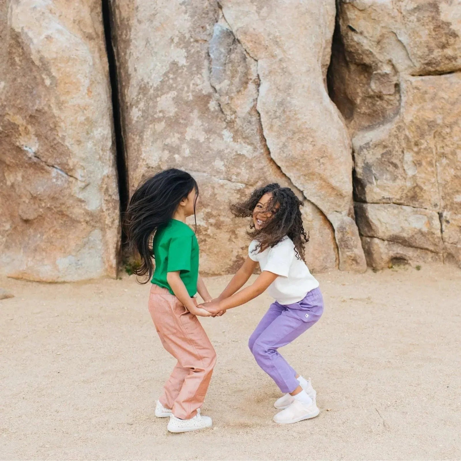 Two kids wearing colorful Jackalo sustainable clothes play outdoors by large rocks.