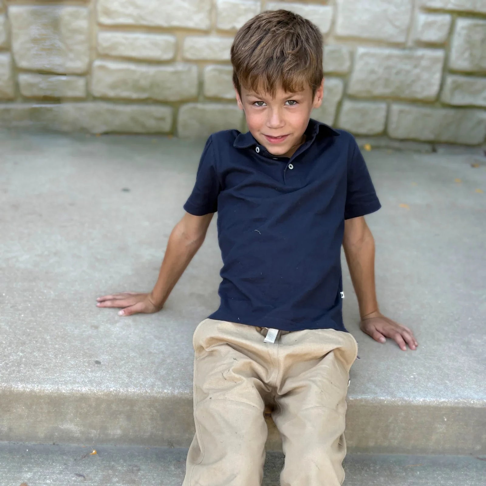 Boy in navy polo and khaki pants from Jackalo sitting on stone steps outdoors