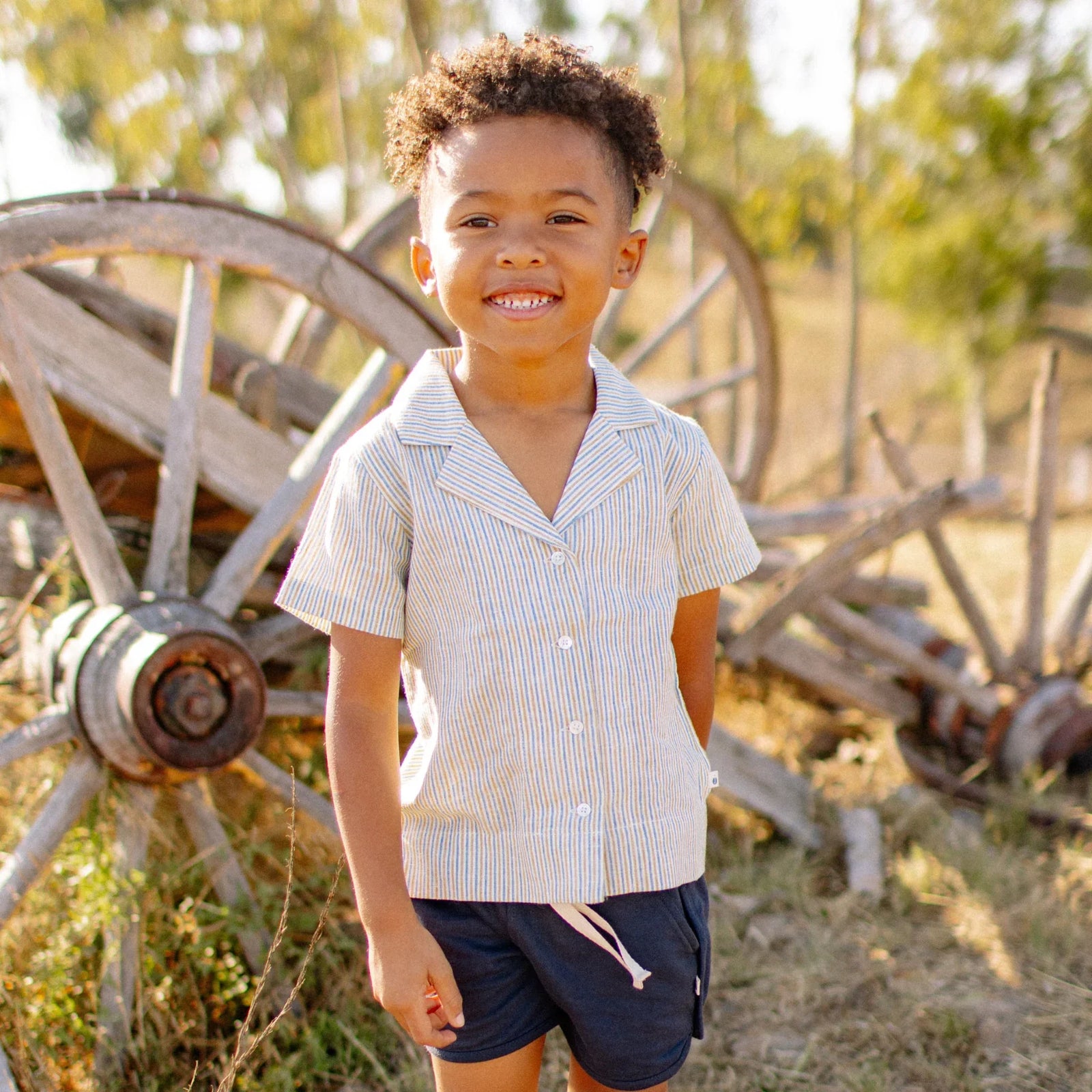 Smiling child in Jackalo camp shirt and shorts outdoors by wooden wagon wheels