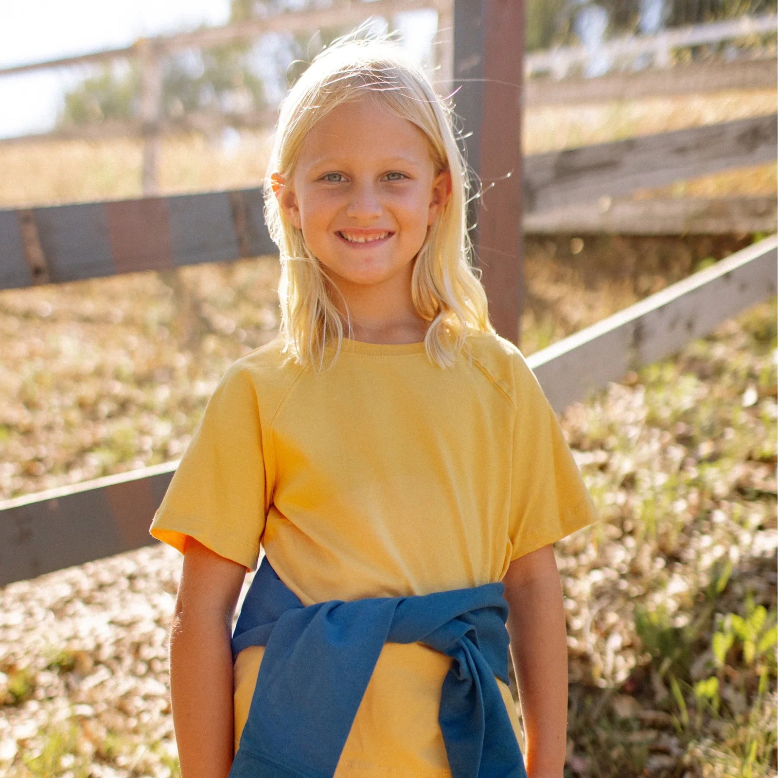Smiling child in Jackalo sustainable yellow shirt outdoors near a wooden fence