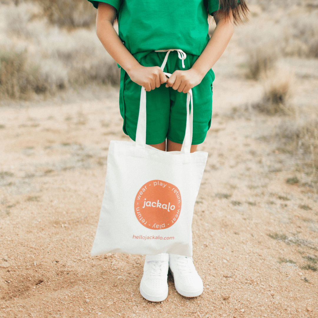 Child in green outfit holding Jackalo organic tote bag outdoors on dirt path