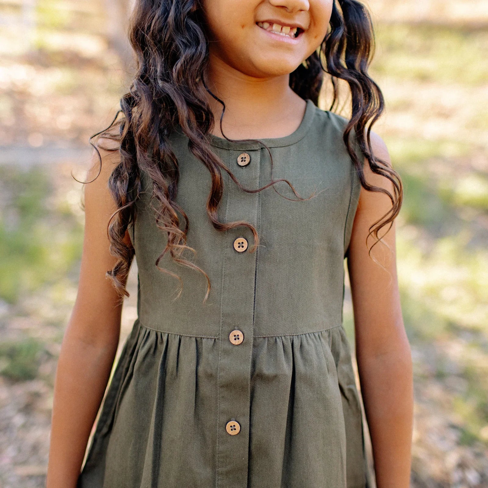 Smiling child wearing olive green sleeveless button-front dress by Jackalo outdoors