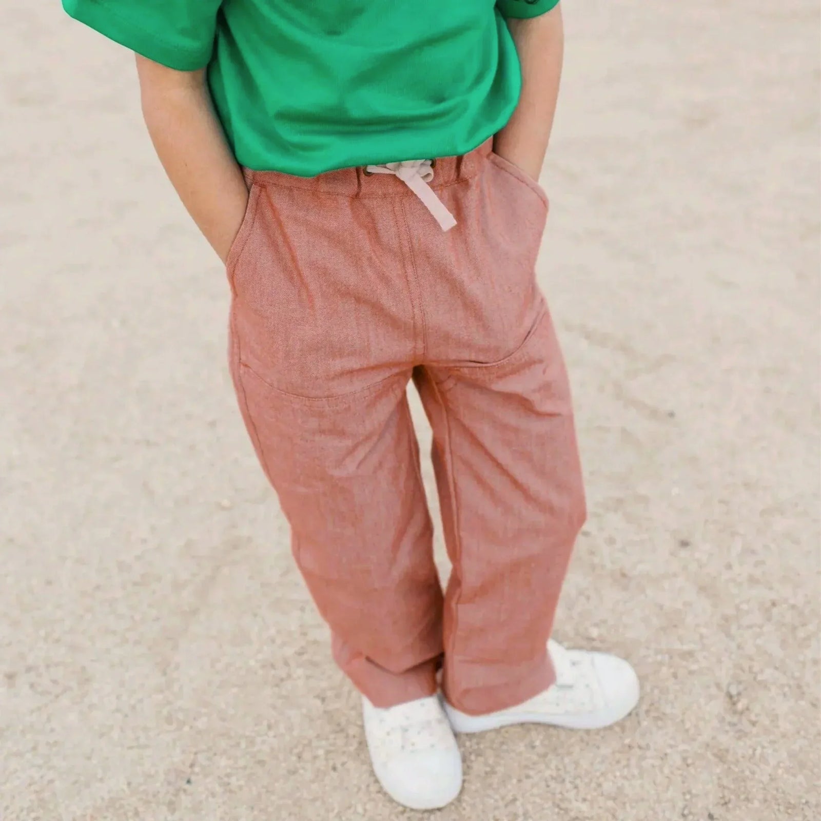 Child wearing Jackalo Jules Pants in chambray, green shirt, and white sneakers on sand