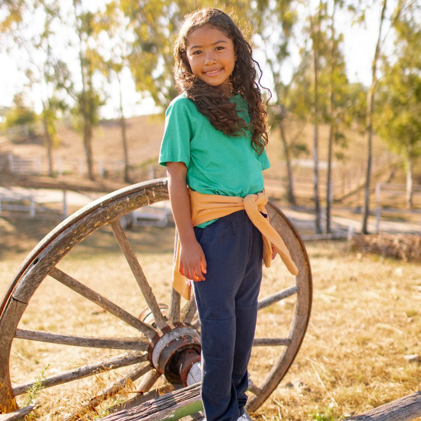 Smiling girl wearing sustainable Jackalo kids clothes stands by rustic wagon wheel outdoors.