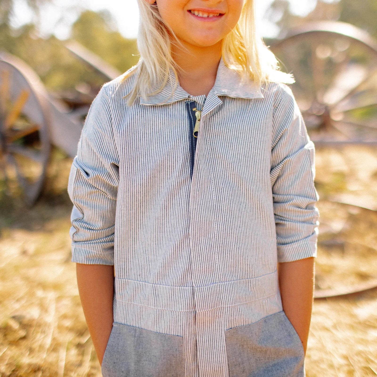 Smiling child wearing striped sustainable Jackalo coverall outdoors with wagon wheels in background