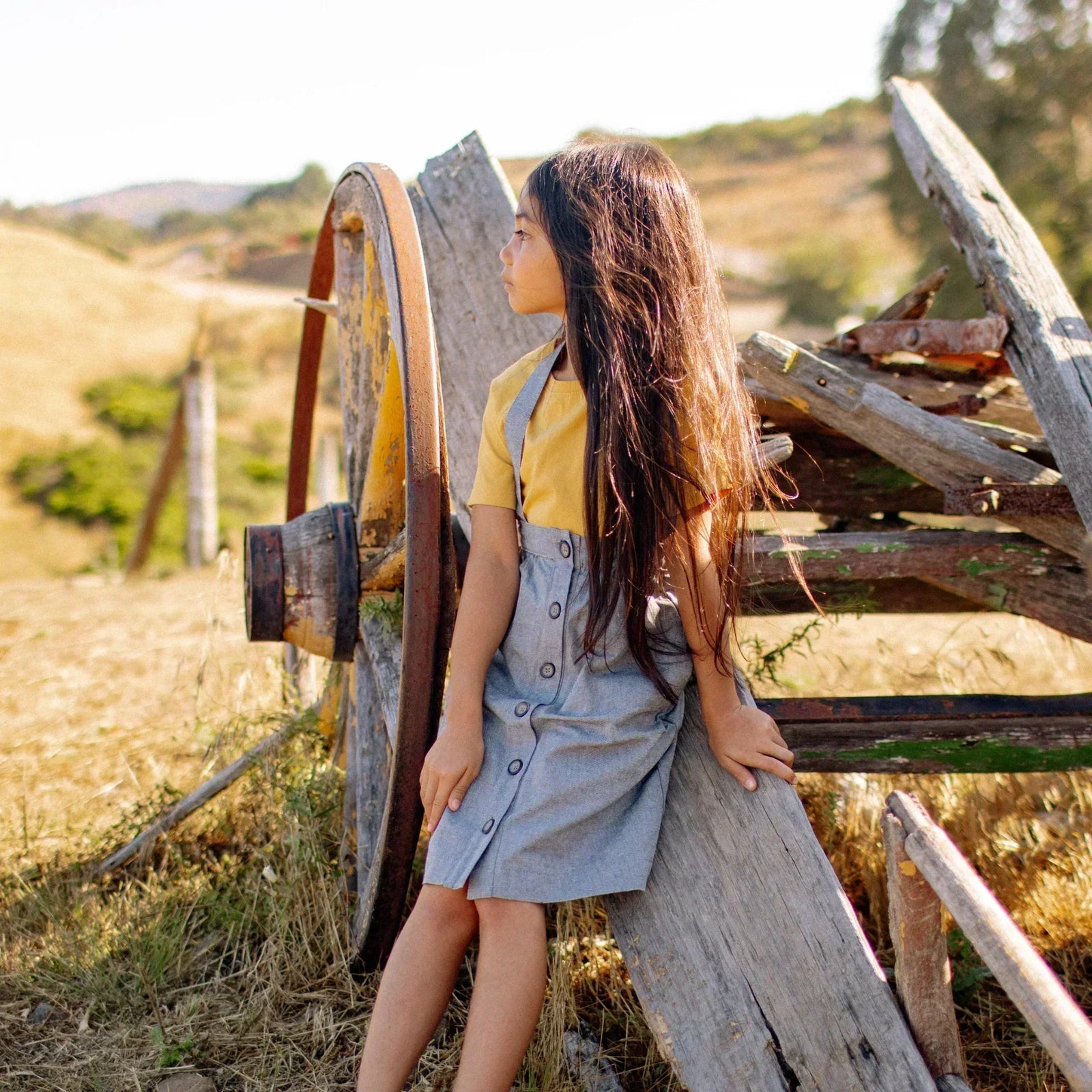 Girl in Jackalo Parker skirt and yellow shirt outdoors by rustic wagon wheel