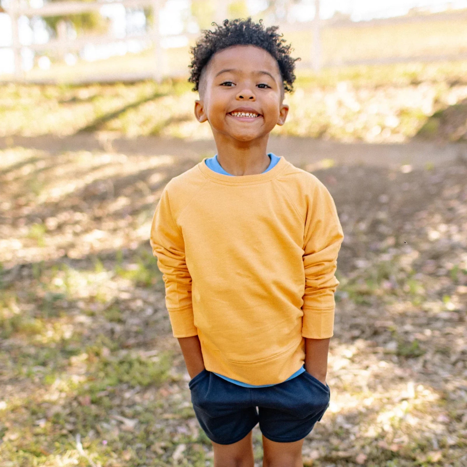 Smiling child wearing a yellow Jackalo sweatshirt and navy shorts outdoors in sunlight