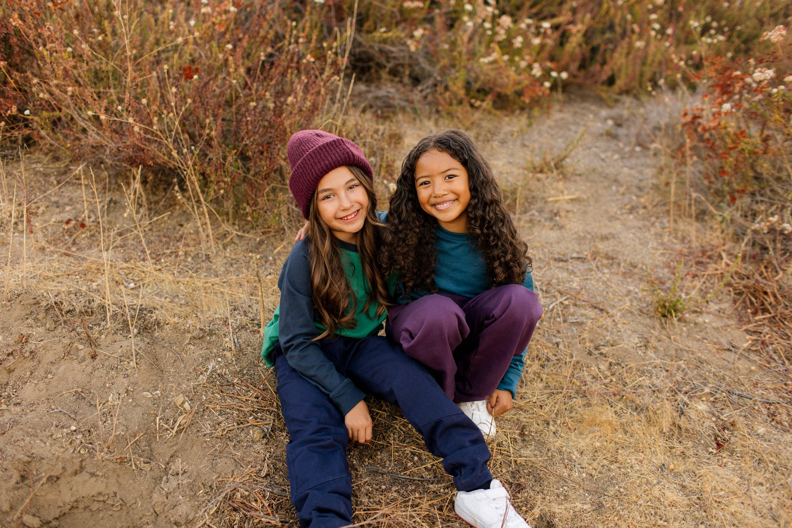 Two kids in cozy, sustainable activewear sitting outdoors, smiling with nature backdrop