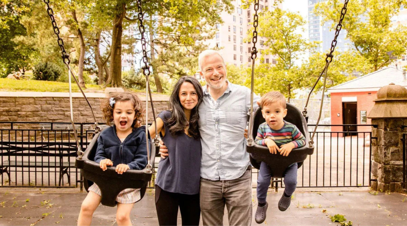 Happy family with two kids in swings at a city park, wearing Jackalo sustainable kids clothes