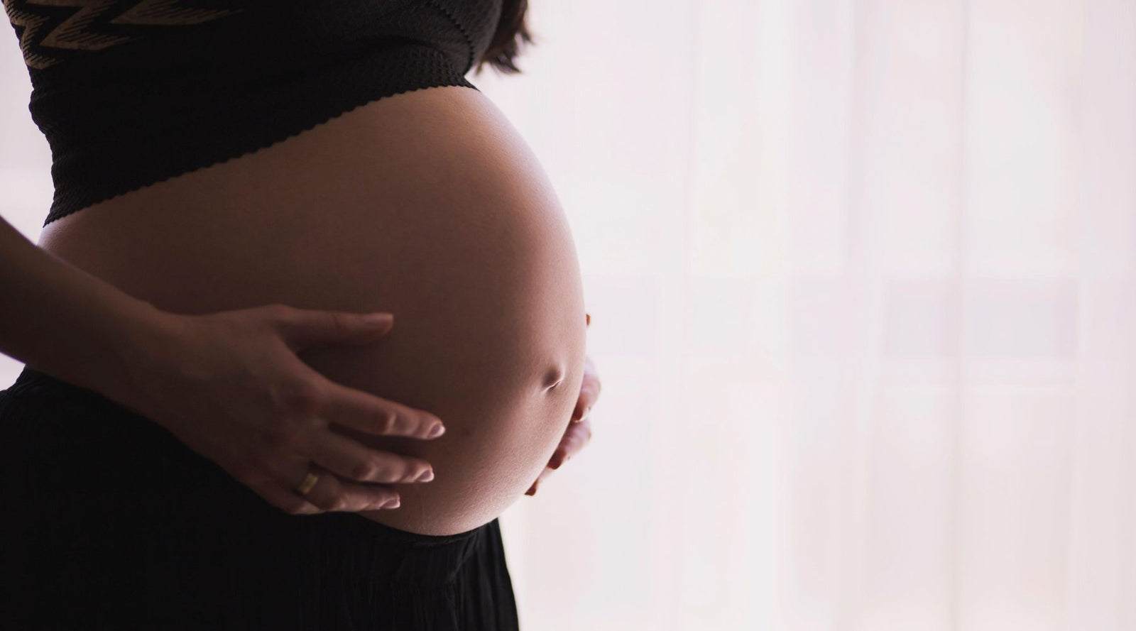 Pregnant woman in black top resting hands on belly, soft natural light background, Jackalo family