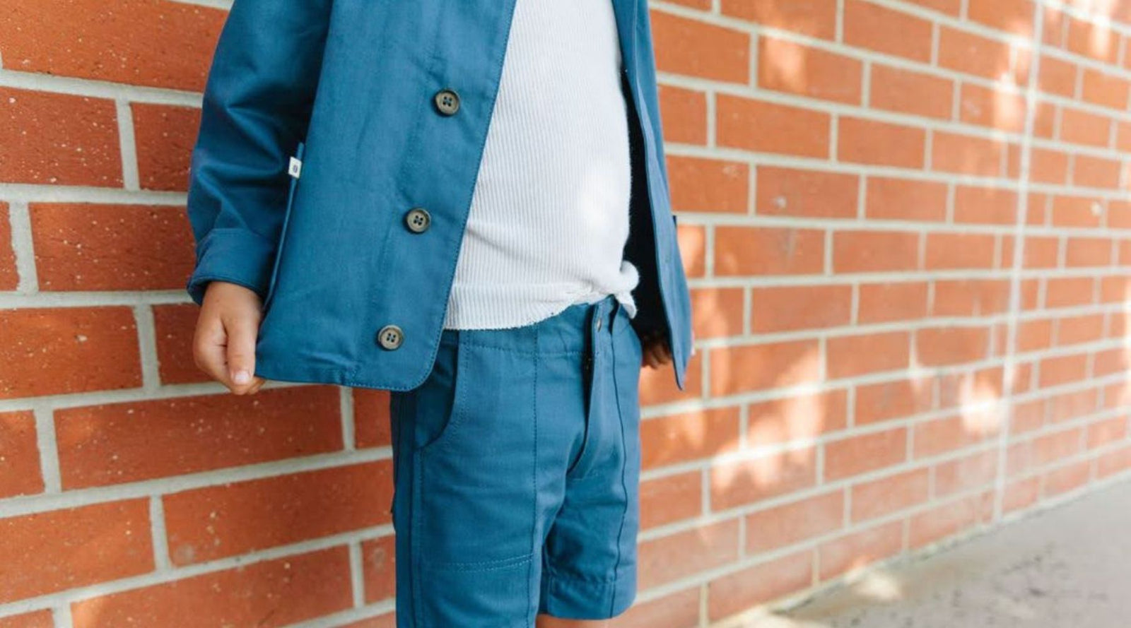 Boy in sustainable blue jacket and shorts with white shirt by Jackalo, standing by brick wall