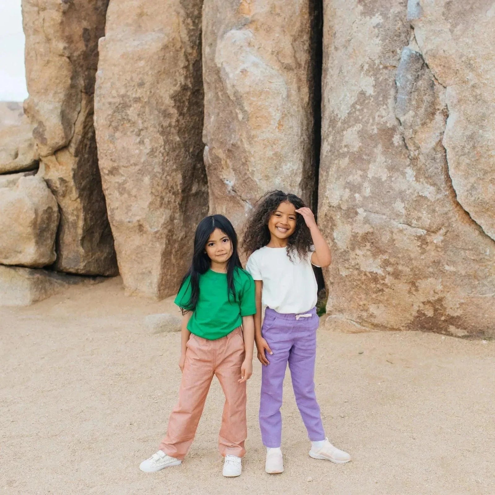 Two kids wearing Jackalo sustainable pants and shirts, smiling outdoors by large rocks.
