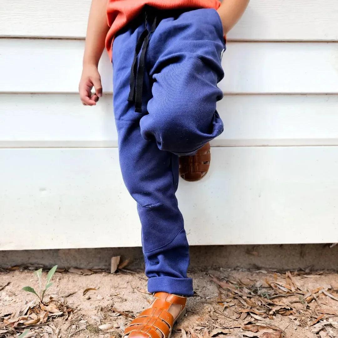 Child wearing blue Jackalo sustainable joggers and sandals, leaning against a white wall