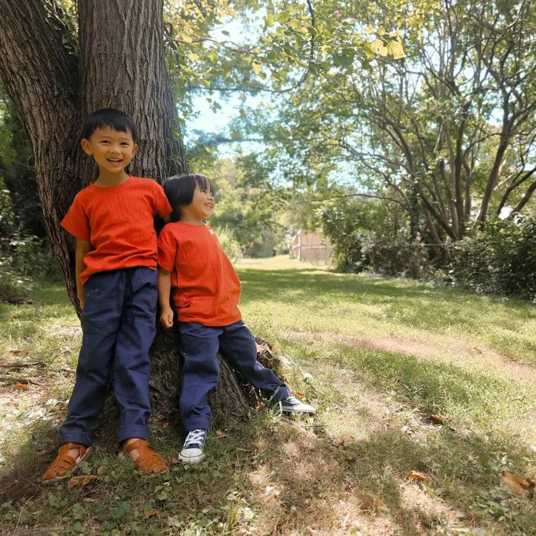 Two kids in Jackalo sustainable red shirts and blue pants smiling outdoors under a tree