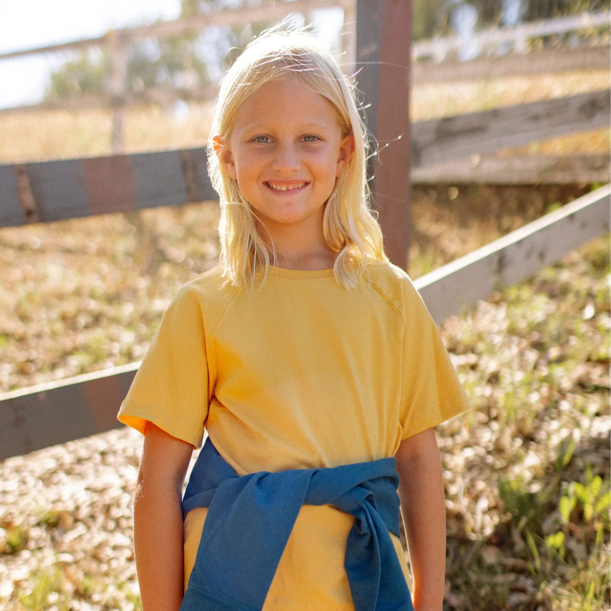 Smiling child in Jackalo sustainable yellow shirt outdoors near a wooden fence