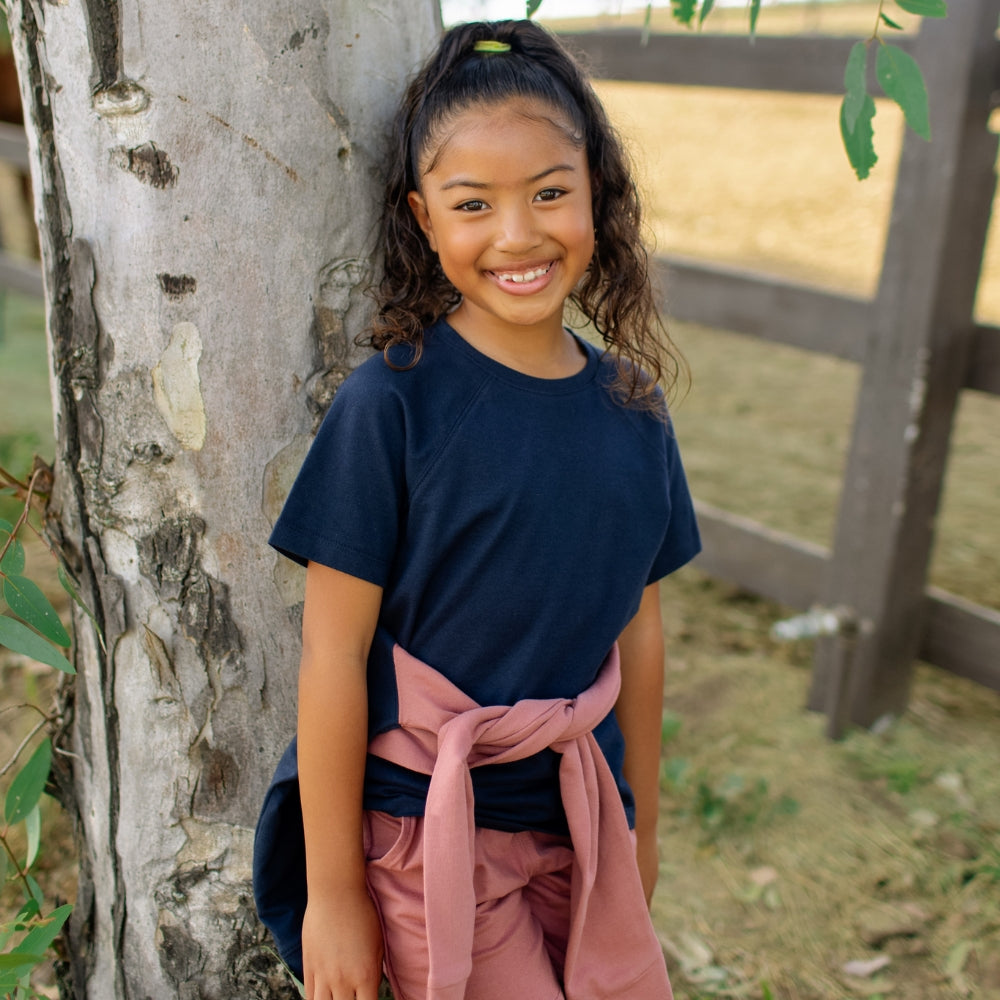 Young girl in a navy shirt and pink shorts standing next to a tree