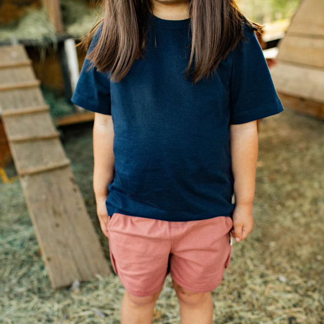 Child wearing a organic navy blue t-shirt and organic pink shorts standing in an outdoor setting with wooden structures.