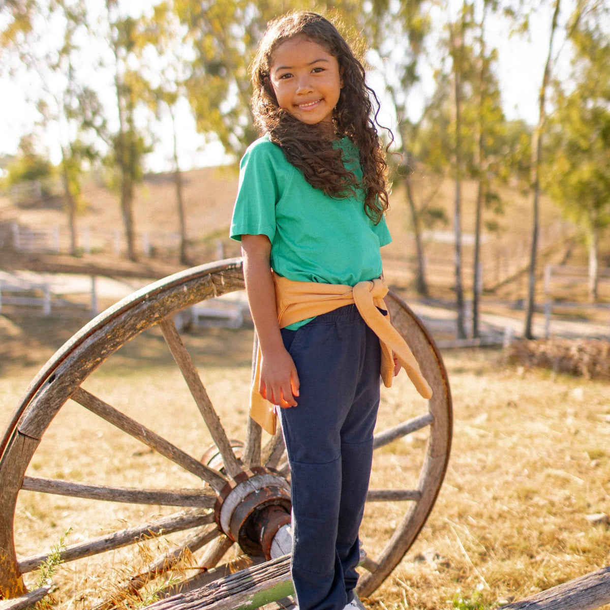Smiling child in Jackalo sustainable kids clothing, navy pants and green tee outdoors by wagon wheel
