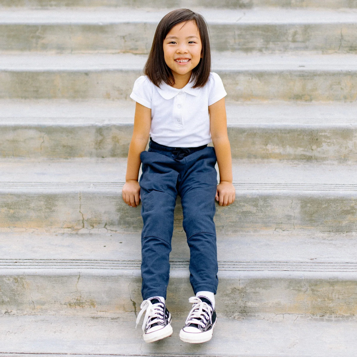 Smiling child in Jackalo sustainable navy pants and white shirt sitting on outdoor steps