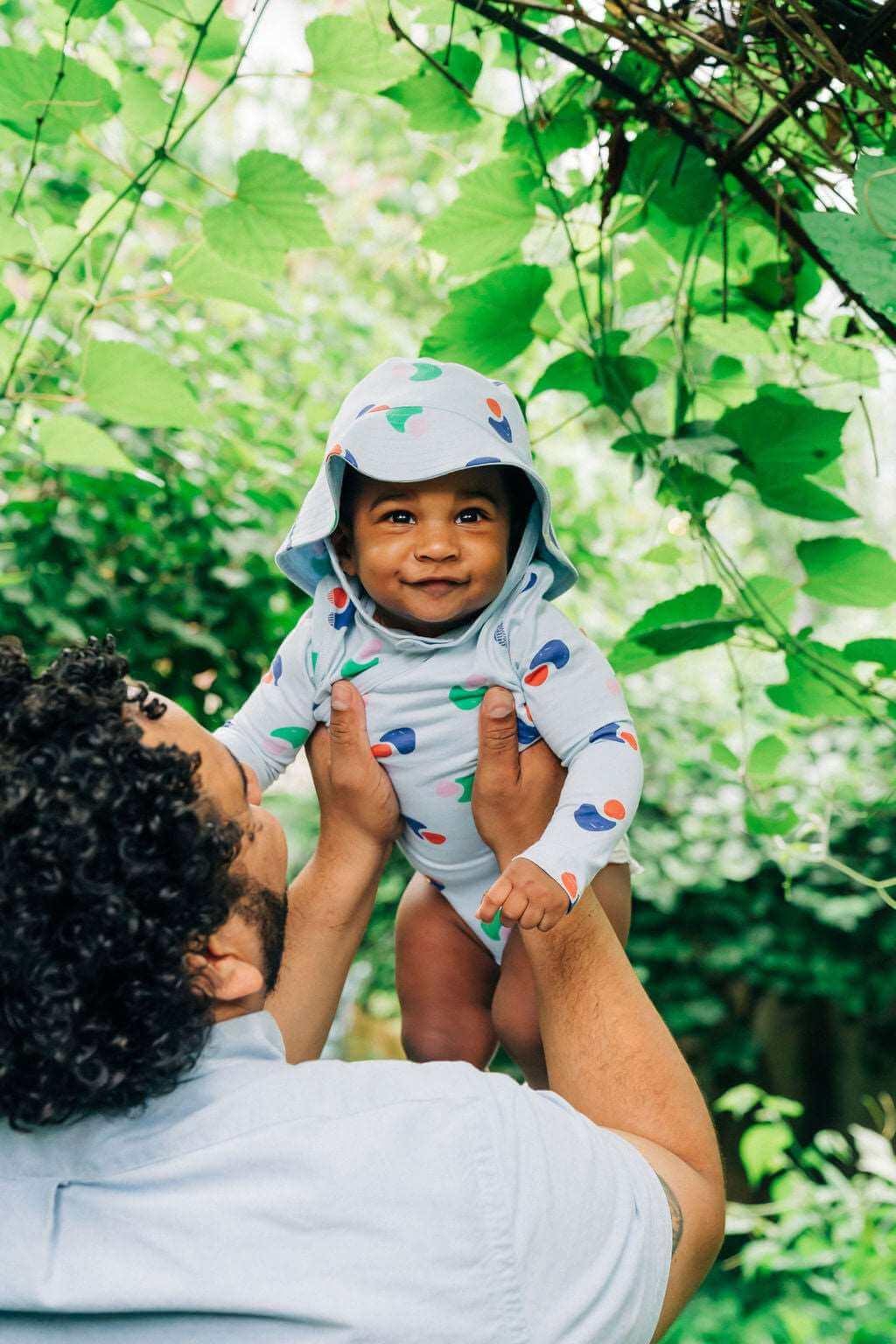 Adult lifting smiling baby in sustainable Jackalo kids clothing outdoors amid greenery