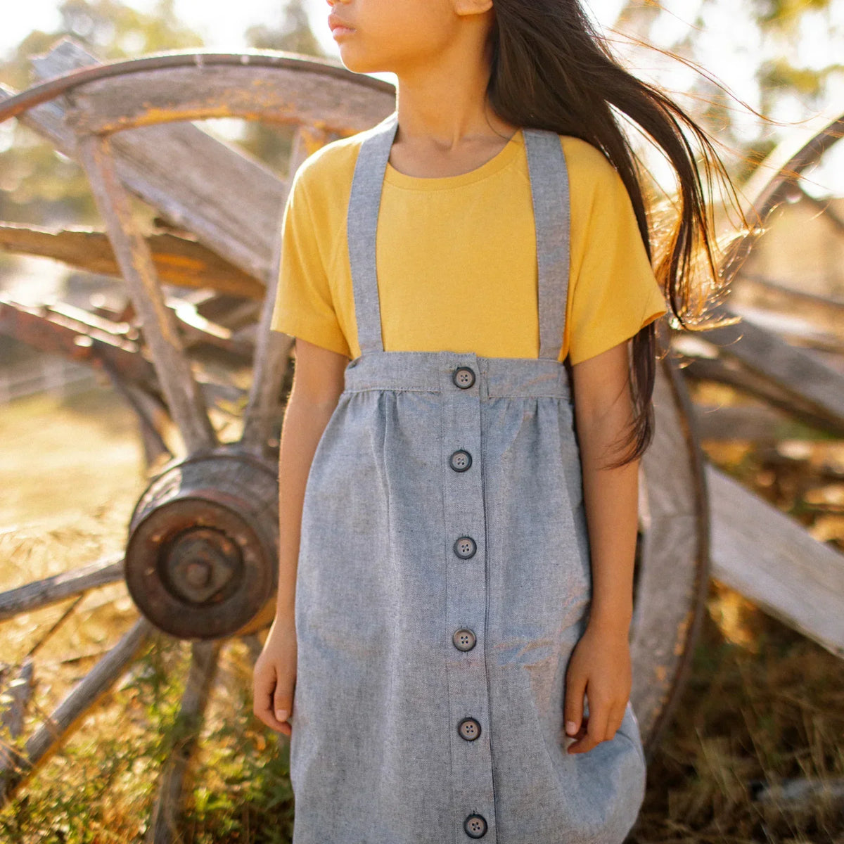 Young girl in sustainable Jackalo denim suspender skirt and yellow shirt outdoors by wagon wheel