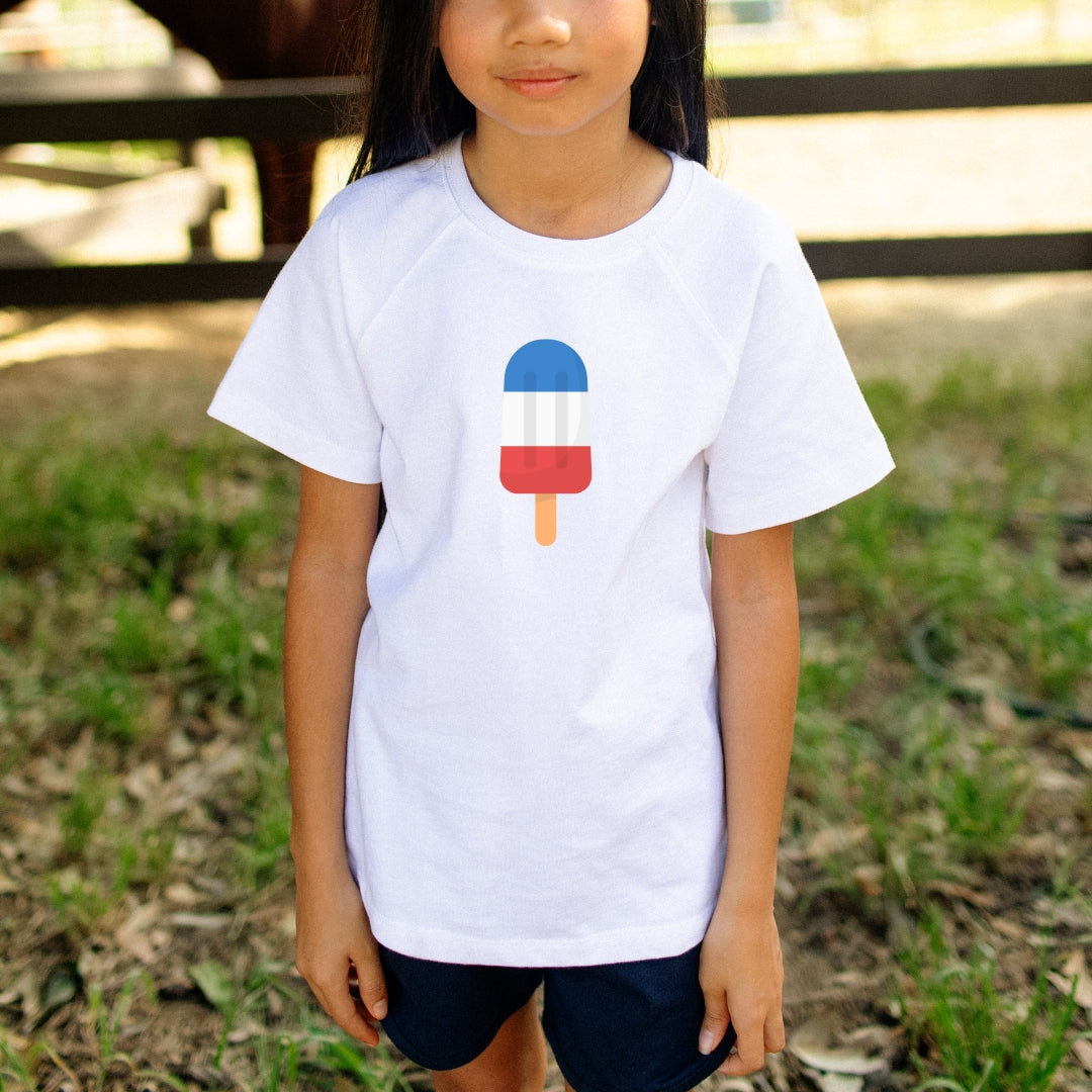 Child wearing a white t-shirt with a red, white, and blue popsicle design outdoors.