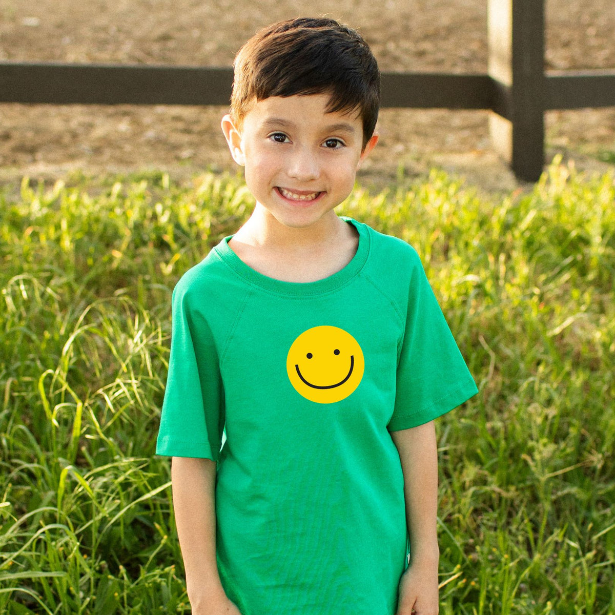 Child wearing a green t-shirt with a yellow smiley face in a grassy field