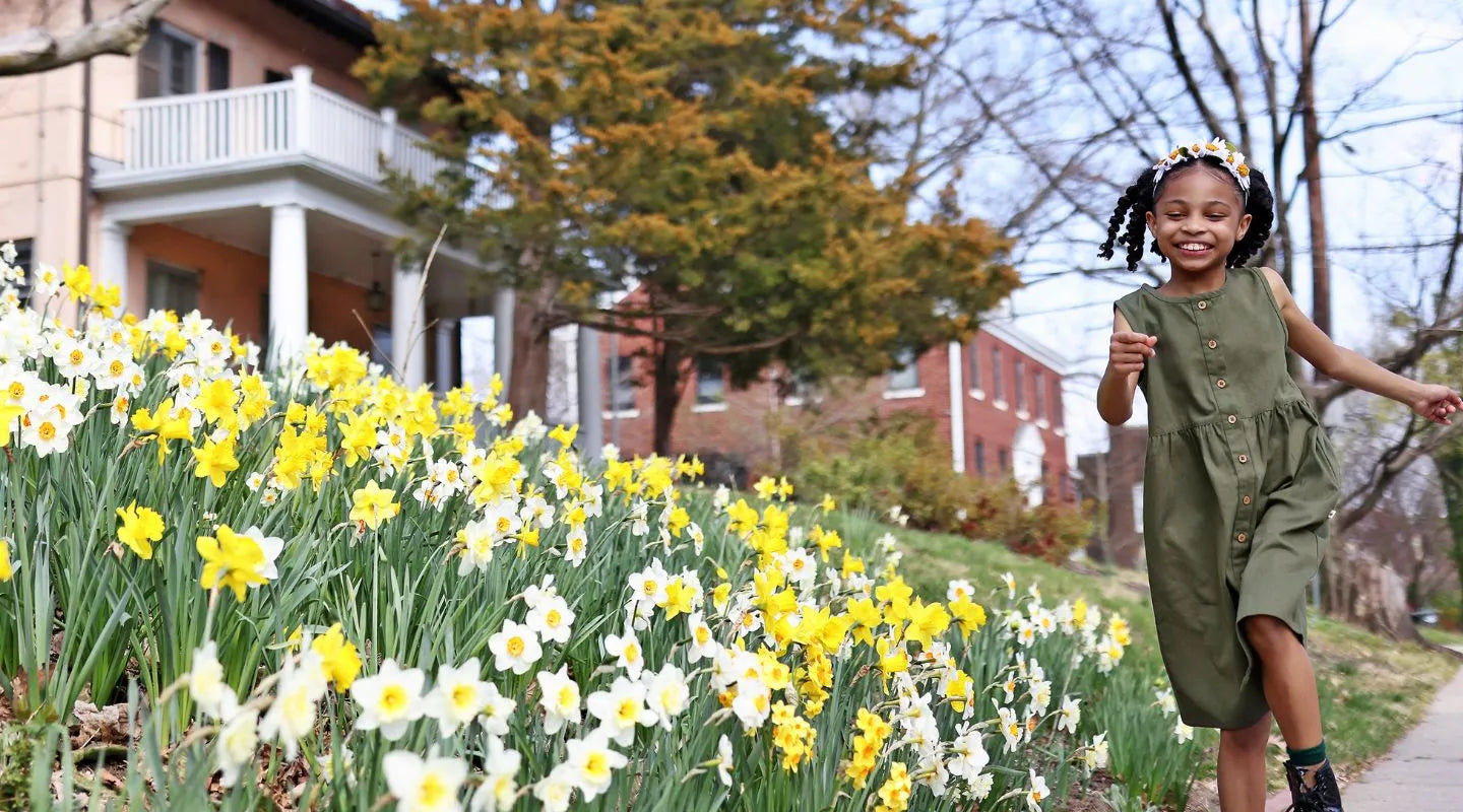 A cheerful girl in a green dress walks down a sidewalk, surrounded by blooming daffodils