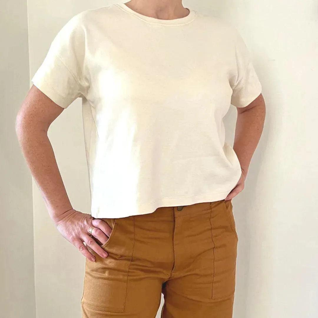 Adult wearing sustainable cream t-shirt and brown pants by Jackalo, standing against white wall