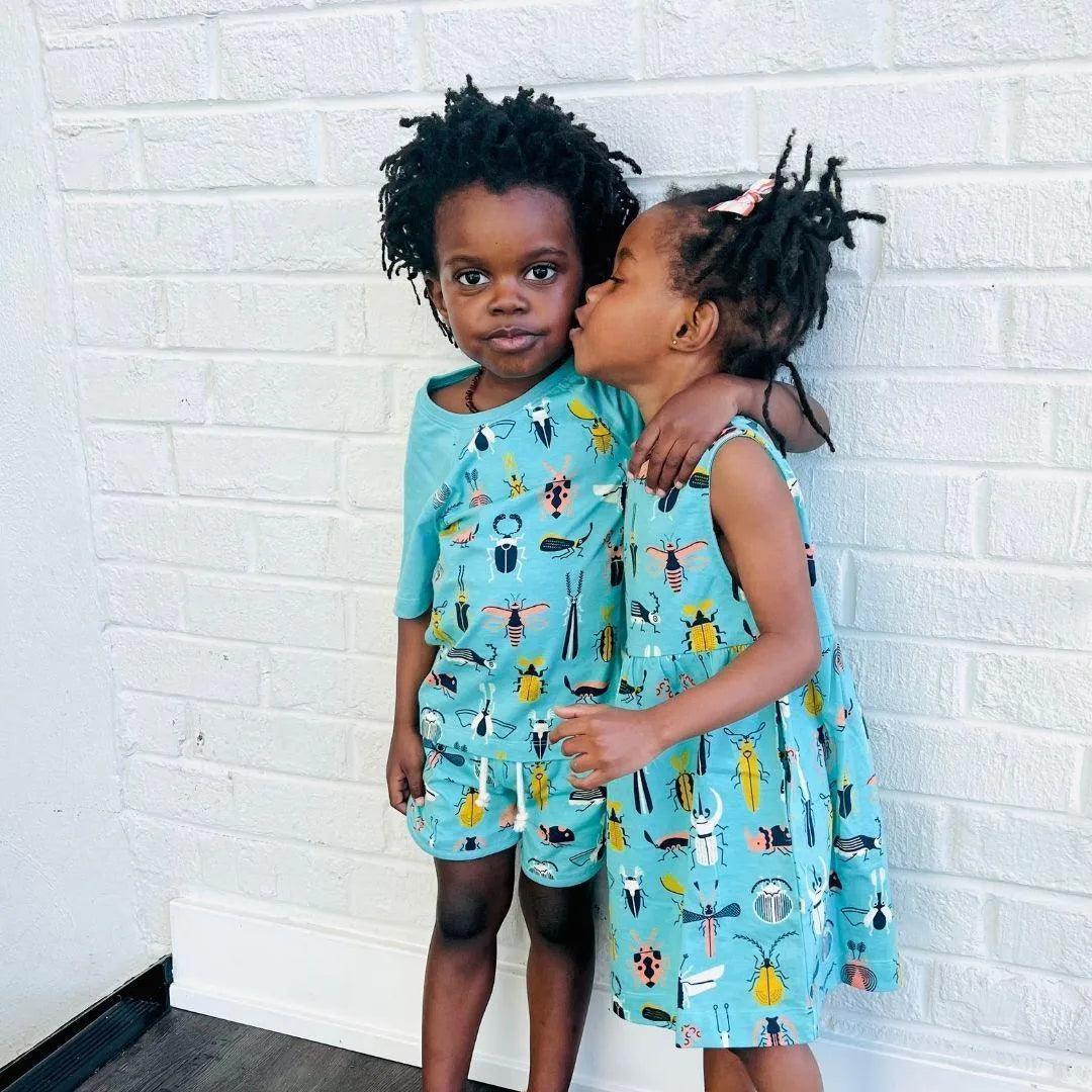 Two kids in matching blue Jackalo beetle print outfits standing by a white brick wall.