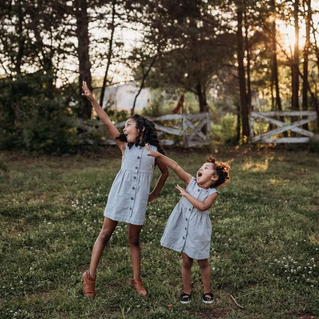 Two girls wearing Jackalo engineer stripe dresses playing outside in a grassy, sunlit yard.