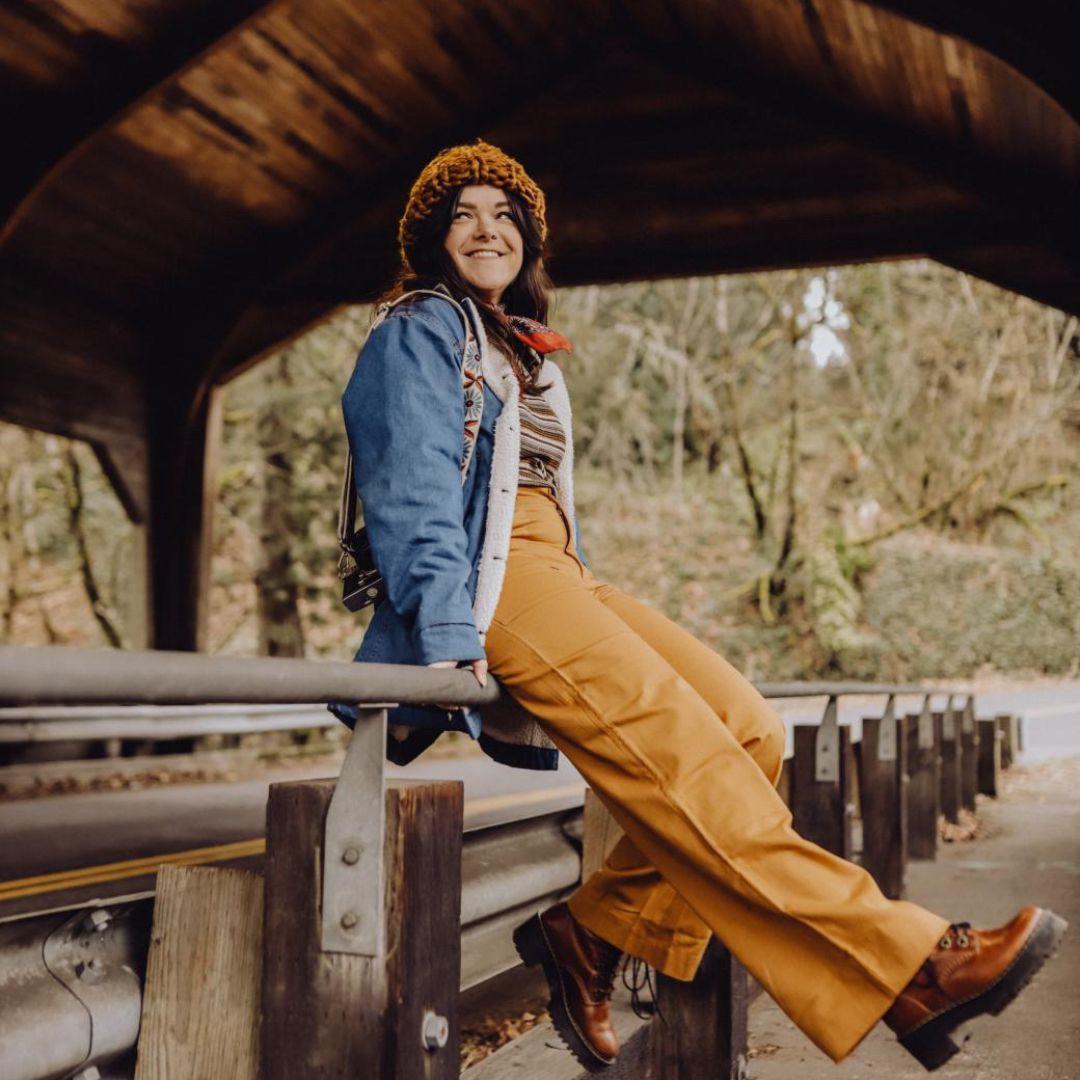 Woman in acorn brown Jackalo Charlie pants and denim jacket smiling on a guardrail outdoors