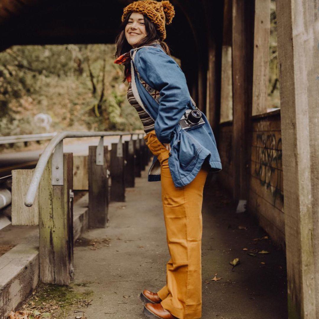 Woman wearing Jackalo Charlie pants in acorn, denim jacket, knit hat, outdoors on a bridge